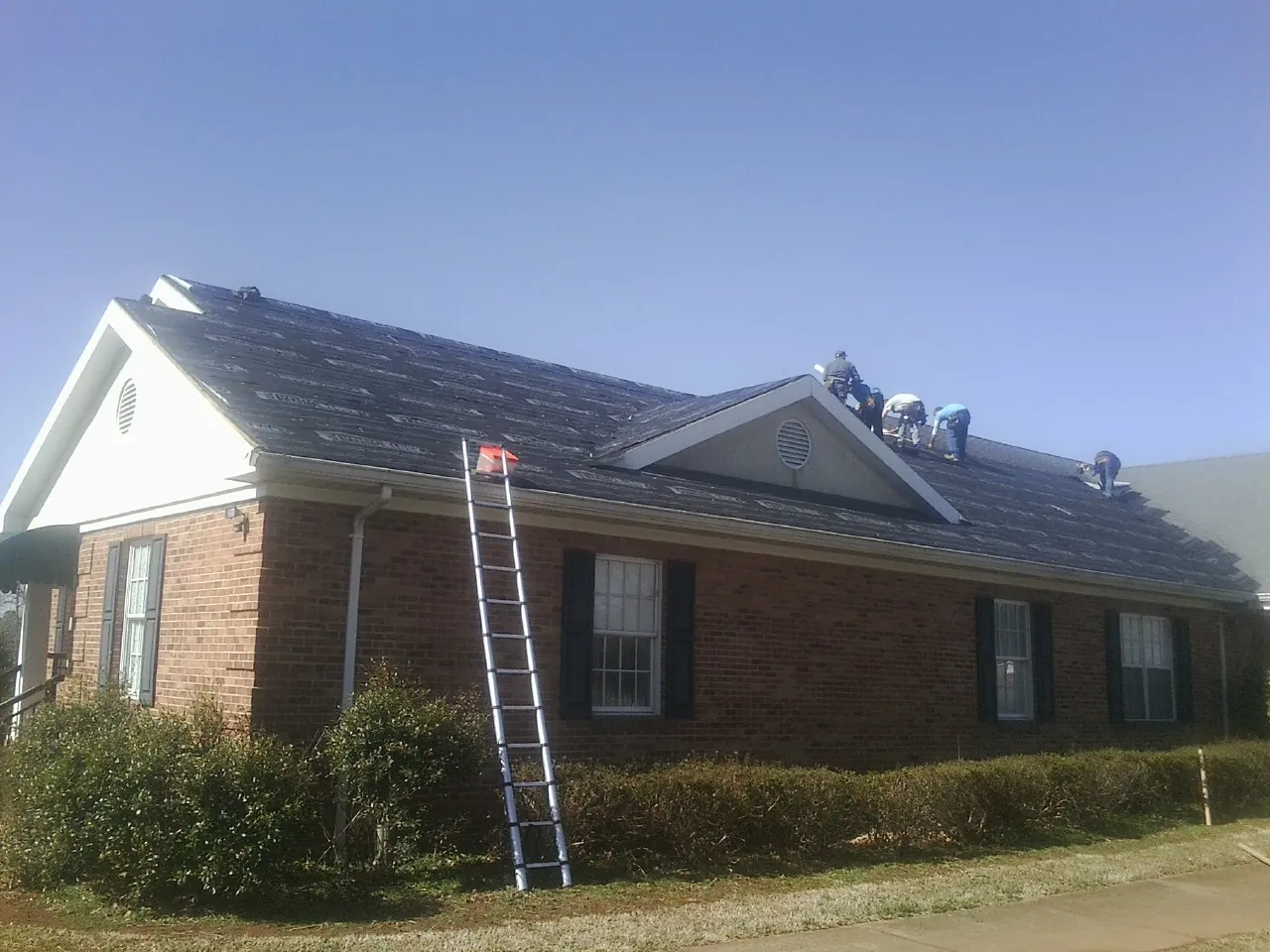 Crew tearing shingles off a brick ranch roof with a ladder set — Emergency Roof Repair work by our crew in Winnie