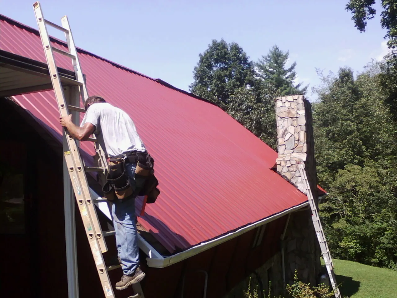 Roofer on a ladder beside a red metal roof and stone chimney — Soffit & Fascia Repair by Roofing Brothers in Winnie